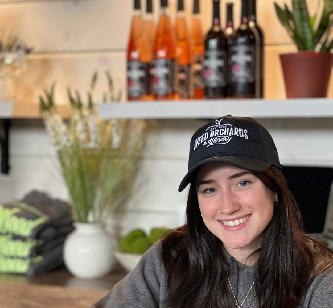 A female leans on the bar with a Weed's Winery baseball hat and wine in the background